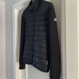 Moncler cardigan  - Snygg svart quiltad jacka från Moncler med dragkedja och långa ärmar. Jackan har en stilren design med Moncler-logga på ärmen och ribbade detaljer vid kragen. Perfekt för våren och hösten