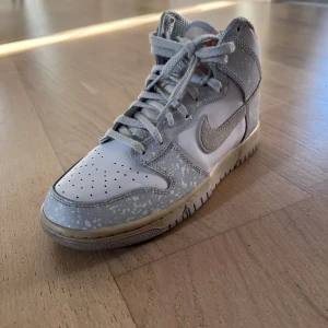 Nike Dunk High i grått och vitt spray limited edition  - Säljer ett par Nike Dunk High sneakers i grått och vitt med ett unikt mönster. Skorna har snörning och en klassisk Nike-logga på sidan. Perfekta för dig som vill ha en stilren och modern look. Man kan få med Chris skydd om man lägger till 50kr 