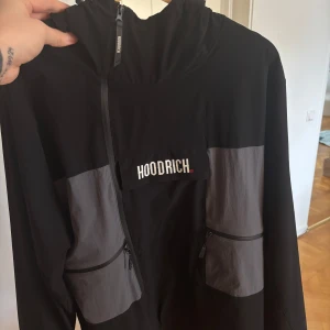 Svart/grå jacka från Hoodrich - Säljer en svart och grå jacka från Hoodrich med huva och dragkedja. Jackan har två stora fickor framtill med dragkedjor och två dolda mindre fickor på bröstet och en på armen. 