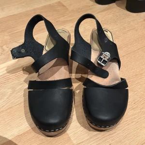 Svarta träskor med klack - Snygga svarta träskor med klack och justerbart spänne. Skorna har en robust träklack. Perfekta för en stilren look.