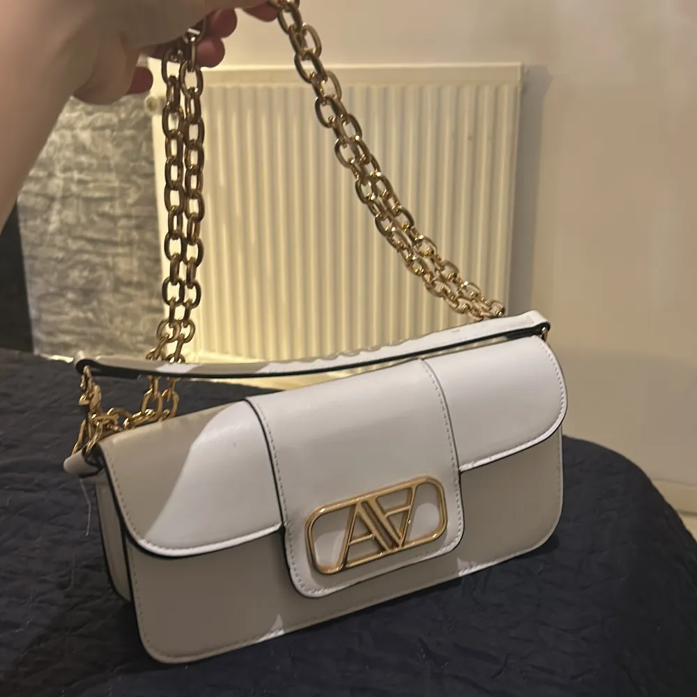 Snygg vit axelväska med elegant guldkedja och stilren design. Väskan har ett dekorativt guldfärgat spänne på framsidan och en praktisk axelrem. Perfekt för att ge din outfit en lyxig touch.. Laukut & Käsilaukut.