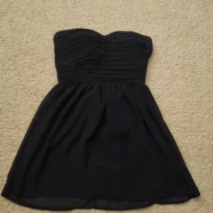 Mörkblå axelbandslös klänning - Snygg svart axelbandslös klänning med dragkedja bak. Den har en elegant rynkad detalj på överdelen och en kort kjol som ger en stilren look.