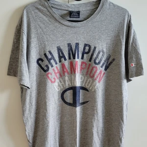Grå t-shirt från Champion - Säljer en grå t-shirt från Champion med tryck i blått och rött. Den har en klassisk rund hals och korta ärmar. Perfekt för en avslappnad stil. 🏆