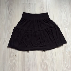 Svart kjol med volang - Säljer en svart kortkjol med volangdetaljer. Kjolen har en elastisk midja för en bekväm passform. Perfekt för att styla med olika toppar och accessoarer.