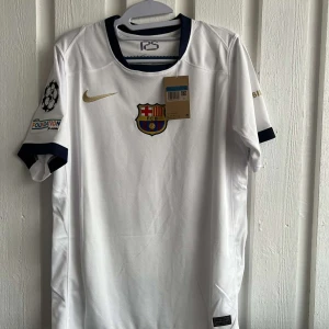 Vit FC Barcelona fotbollströja - Säljer en vit FC Barcelona fotbollströja från Nike med klubbens emblem på bröstet och Lamine Yamal 19 tryckt på ryggen. Tröjan har korta ärmar och detaljer i guld och svart. Perfekt för fans av klubben!