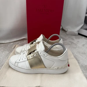 Valentino Garavani sneakers i vitt och guld - Snygga Valentino Garavani sneakers i vitt med en bred gulddetalj över snörningen. Skorna har en klassisk design med rund tå och en bekväm sula. Använda men i bra skick, tar emot prisförslag, skriv om ni har några frågor!❣️❣️