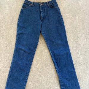Blå jeans från Wrangler - Säljer ett par klassiska blå jeans från Wrangler. De har en straight passform och är tillverkade i slitstarkt denim. Byxorna har en traditionell femficksdesign och en läderpatch med logotyp på bakfickan.