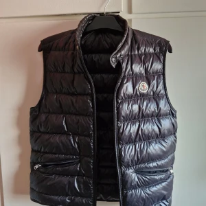 Svart guiväst från Moncler - Snygg svart gui väst från Moncler med dragkedja och två praktiska fickor framtill. Västen har en klassisk design med Monclers logotyp på bröstet. Perfekt för kyligare dagar när du vill hålla stilen. Jag säljer denna för att den var förlåten för mig och inte används. Den är i fint skick och helt äkta med en fungerande NFC tag. Kvitto och allt i bilden följer med vi köp. !!Pris kan diskuteras vid snabb affär!!