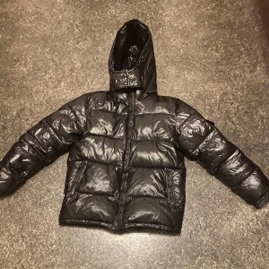 Svart pufferjacka från Moncler - Snygg svart pufferjacka från Moncler med huva och dragkedja. Jackan har långa ärmar och en glansig finish som ger en modern look. Perfekt för kyliga dagar.