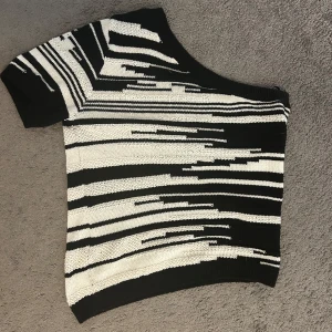 Svartvit offshoulder stickad topp - Unik stickad topp i svart och vitt med coolt randigt mönster och asymmetrisk offshoulder-skärning. Kortärmad och loose fit, perfekt för dig som vill sticka ut med en edgy vibe. Mjuk och bekväm, passar till både jeans och kjol.