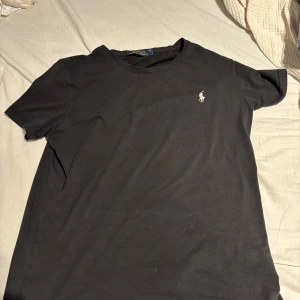 Svart t-shirt från Polo Ralph Lauren - Svart t-shirt från Polo Ralph Lauren med klassisk passform och korta ärmar. Ikoniska broderade loggan i vitt på bröstet. Tillverkad i mjuk bomull som känns skön mot huden. Enkel och stilren design som funkar till allt.