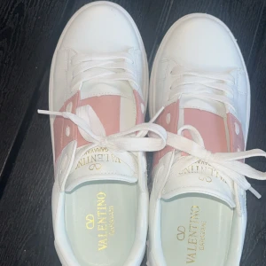 Valentino Garavani sneakers vit/rosa - Snygga Valentino Garavani sneakers i vitt skinn med bred rosa panel och coola nitar på sulan. Klassisk rund tå, platt sula och vita snören. Perfekta för dig som vill ha en stilren men ändå unik look.
