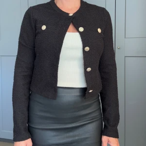 Svart cardigan jacka från H&M med guldknappar - Stilren svart cardigan jacka från H&M med snygg struktur och dekorativa guldknappar framtill. Jackan är croppad, har rund halsringning och långa ärmar. Perfekt för dig som vill ha en klassisk men trendig look.