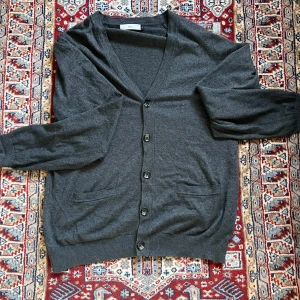 Mörkgrå kofta från Bläck, storlek L - Mysig mörkgrå kofta från Bläck i storlek L. Klassisk v-ringning, knäppning med fem knappar och två fickor framtill. Långärmad och i mjukt material som passar perfekt till lager-på-lager. Enkel och stilren design som funkar till det mesta.