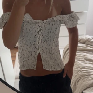 Vit blommig blus med volangärmar - Supersöt vit blus med svart blommigt mönster och volangiga axelband ifrån H&M . Toppen är croppad, har knappar framtill och kan bäras offshoulder eller på axlarna.🩷