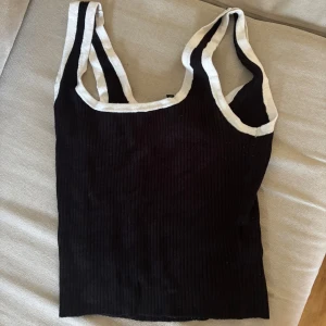 Svart ribbad linnetopp från h&m  - Säljer en svart ribbad linnetopp från h&m med vita kontrastkanter runt hals och axelband. Toppen är ärmlös och har en tight passform som sitter snyggt på kroppen. Storlek M 