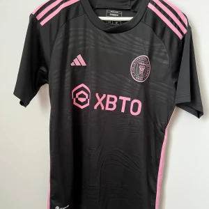 Inter Miami Messi fotbollströja svart/rosa - Inter Miami CF fotbollströja med Messi 10 på ryggen. Svart bas med rosa detaljer, klubbmärke och sponsortryck framtill. Adidas-logga och klassiska ränder på axlarna. Tillverkad i lätt och ventilerande funktionsmaterial.