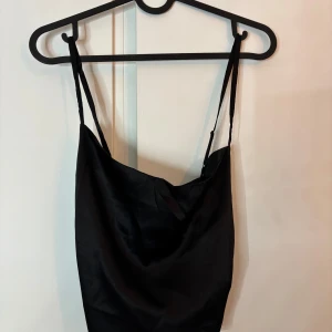 Svart satin linnetopp från NA-KD - Snygg svart linnetopp i satin med tunna axelband och vattenfallsringning. Toppen har croppad passform och smockad detalj baktill för extra komfort. Storlek 38. Helt ny med prislapp