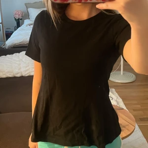 Svart utsvängd t-shirt från Kappahl - En svart basic t-shirt från Kappahl i mjuk bomull. Klassisk rund halsringning och korta ärmar samt utsvängd midja. Perfekt enkel passform som funkar till allt. Snygg att styla med jeans eller shorts för en clean look.