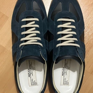 Maison Margiela Replica sneakers blå mocka - Maison Margiela Replica sneakers i mörkblå mocka och skinn med vita snören. Klassisk retrodesign med rund tå och platt sula. Snygga detaljer och stilren look