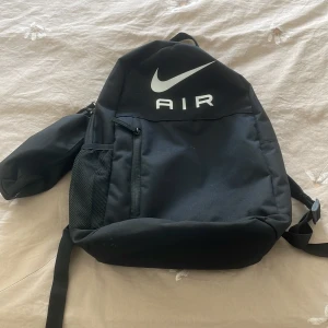 Svart Nike Air ryggsäck med pennfack - Säljer en svart Nike Air ryggsäck med stort vitt logotryck framtill. Ryggsäcken har vadderade axelband, meshficka på sidan och ett separat pennfack som sitter fast på ena sidan. Perfekt för skolan eller gymmet, rymmer allt du behöver.
