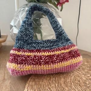 Stickad färgglad handväska - Unik stickad handväska i blått, vinrött, gult och rosa. Väskan har en mjuk, rundad form och ett brett handtag. Perfekt för dig som vill ha en personlig accessoar med retrovibbar. Väskan är handgjord och har ett coolt randigt mönster.