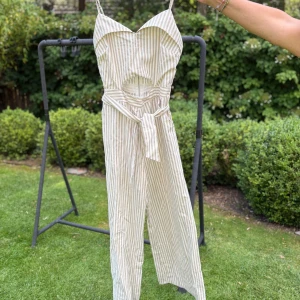 Randig byxdress från TIC:TOC, beige/vit - Snygg randig byxdress från TIC:TOC i beige och vitt med smala axelband, cut-out vid midjan och knytband. Vida ben och dragkedja bak. Perfekt för varma dagar när du vill ha en chill men trendig look.