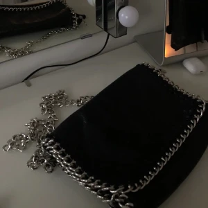 Svart axelväska från TIAMO med kedja - Säljer en svart axelväska från TIAMO i skinn med silverfärgad kedja som axelrem och cool kedjedetalj runt hela väskan. Stilren och edgy design som lyfter vilken outfit som helst. Perfekt storlek för det viktigaste och ger en snygg statement-look. 