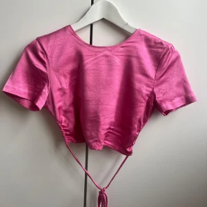 Rosa croppad topp med snörning - Rosa croppad topp med korta ärmar och knytdetaljer i ryggen. Toppen har smala snören som korsas i ryggen och knyts i midjan. Materialet har en lätt glans.