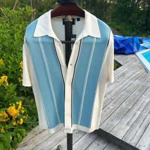 Randig stickad piké Grand Le Mar - Snygg stickad skjorta från Grand Le Mar i storlek Large. Skjortan har korta ärmar, klassisk krage och knappar framtill. Färgen är vit med ljusblå och svarta ränder på framsidan. Perfekt för dig som gillar retrovibbar och stilren design. Aldrig använd. Öppen för prisförslag.