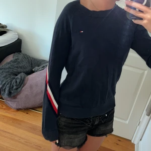 Mörkblå Tommy Hilfiger tröja med ränder - Storlek 164/XS mörkblå långärmad tröja från Tommy Hilfiger med klassiska röda och vita ränder längs ärmarna och över ryggen. Liten broderad logga på bröstet och ribbad halsringning. Skön och lätt tröja men måste tyvärr säljas då den blivit för liten.