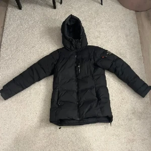 Svart dunjacka Sail Racing med huva - Säljer en svart dunjacka från Sail Racing, modell Antarctica. Jackan har en längre passform, stor huva och dragkedja framtill. Broderad logga på bröstet och patch på ärmen. Perfekt för kalla dagar när du vill hålla värmen och ändå se clean ut.