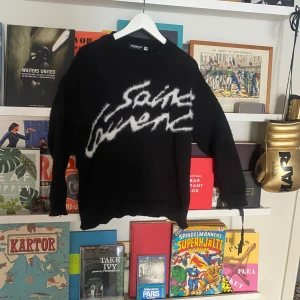 Svart Saint Laurent stickad tröja - Kan gå ner i pris vid SNABB affär! Svart stickad tröja från Yves Saint Laurent med stor vit logga i fuzzy stil över bröstet. Tröjan har rund hals, långa ärmar och en loose fit. Perfekt för dig som vill sticka ut med en ikonisk designertröja och gillar streetwear-vibe.