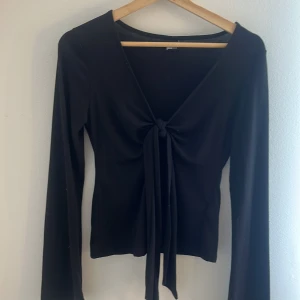 Svart omlottopp från Gina Tricot - Svart långärmad omlottopp från Gina Tricot med snygg knytning framtill och djup v-ringning. Toppens smidiga material gör den bekväm och lätt att styla till olika looks. Perfekt för dig som gillar trendiga och stilrena plagg.