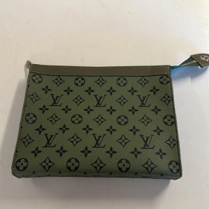 Grön Louis Vuitton kuvertväska i skinn - Snygg grön kuvertväska från Louis Vuitton i skinn med klassiskt monogrammönster i svart. Väskan har silverfärgad dragkedja och röd insida med kortfack. Perfekt för att bära det viktigaste och ge din outfit en lyxig touch.