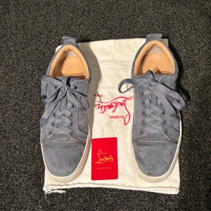 Ljusblåa mocka sneakers från Louboutin - Snygga sneakers från Christian Louboutin i grå mocka med klassiska röda sulor. Skorna har rund tå, platt vit sula och breda snören. Insidan är fodrad i beige läder för extra komfort. Perfekta för dig som vill sticka ut med exklusiva detaljer. Ingen box finns tyvär då den är borttappad, kortet samt dustbag medföljer 