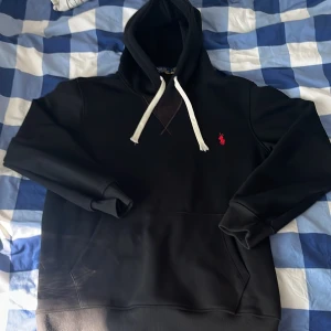 Svart hoodie från Polo Ralph Lauren - Svart hoodie från Polo Ralph Lauren med vit snörning och klassisk röd broderad logga på bröstet. Tröjan har huva, känguruficka och ribbade muddar. Perfekt för en chill och stilren look. Tröjan är i ny skick eftersom jag inte använde den för den ej passa. Storlek M men sitter lite mer som S.