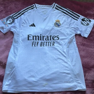 Real Madrid Mbappé 9 matchtröja Adidas - Säljer en vit Real Madrid fotbollströja från Adidas med Mbappé och nummer 9 på ryggen. Tröjan har svarta detaljer, klubbmärke, Champions League-märke, HP-logga på ärmen och 'Emirates Fly Better' på bröstet. Tillverkad i lätt funktionsmaterial (AEROREADY).