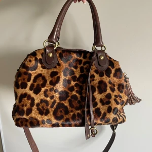 Leopardmönstrad handväska  - Cool handväska i leopardmönstrad pälsimitation med bruna skinnhandtag och axelrem. Väskan har guldfärgade metalldetaljer, dragkedja och en snygg tofs som accessoar. Rymligt foder med innerfack och dragkedja. Perfekt för dig som älskar leopard!💗💗