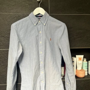 Ljusblå Ralph Lauren Skjorta - Klassisk ljusblå skjorta från Polo Ralph Lauren i slim fit-modell. Skjortan har button down-krage, långa ärmar och broderad logga på bröstet. Tillverkad i bomull och stängs med vita knappar framtill. Perfekt för en clean och stilren look.