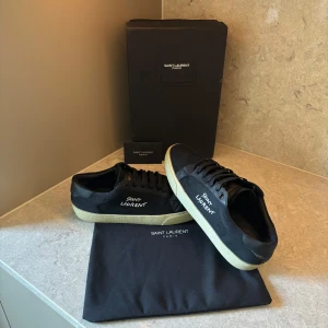 Svarta Saint Laurent sneakers - Snygga svarta sneakers från Saint Laurent med vit logga på sidan. Skorna har en klassisk låg profil, svarta snören och vit sula. Ovandelen är i canvas med detaljer i skinn, vilket ger en stilren och exklusiv känsla. Perfekta för dig som gillar minimalistisk design med lyxig touch.