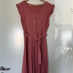 Zauberstern rosa klänning, storlek M - Rosa klänning från Zauberstern i storlek M, med v-ringning och dekorativa knappar framtill. Klänningen har korta, volangprydda ärmar och ett avtagbart band i midjan för att framhäva figuren. Perfekt för en somrig och avslappnad look. Nyskick. 100% viskos.   Mått: Längd ca 105 cm Byst ca 53 cm Midja ca 52 cm Axel till axel ca 42 cm