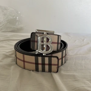 Rutig Burberry bälte med B-spänne - Snyggt bälte från Burberry med klassiskt rutigt mönster i beige, svart och rött. Bältet har ett stort silvrigt B-spänne och är gjort i skinn. På bältet syns även Burberrys ikoniska riddarlogga. Perfekt accessoar för att lyfta din outfit.