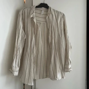  skjorta i linne - Snygg beige skjorta med tunna svarta ränder, tillverkad i ett luftigt linnematerial. Skjortan har lång ärm, klassisk krage och knäppning framtill. Perfekt för en avslappnad och stilren look.