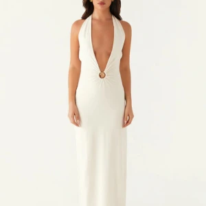 Vit långklänning med djup ringning - Supersnygg vit långklänning med djup v-ringning och halterneck. Klänningen har en dekorativ ringdetalj framtill, öppen rygg och knytband i nacken och ryggen. Materialet är mjukt och följsamt, perfekt för sommarkvällar eller speciella tillfällen.