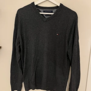 Mörkgrå v-ringad tröja Tommy Hilfiger - Mörkgrå v-ringad tröja från Tommy Hilfiger med liten broderad logga på bröstet. Tröjan är långärmad och har ribbade muddar vid ärmslut och nederkant. Perfekt för lager-på-lager och stilren look.