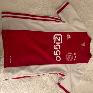 Ajax fotbollströja Adidas röd/vit - Ajax Amsterdam matchtröja från Adidas i klassisk röd och vit design. Tröjan har korta ärmar, klubbmärke och Ziggo-logga på bröstet samt tre stjärnor ovanför klubbmärket. Tillverkad i lätt och ventilerande Aeroready-polyester.