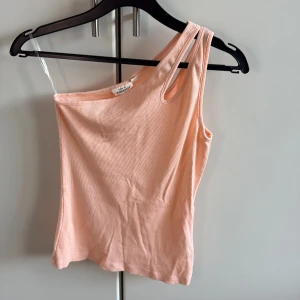 Ljusrosa ribbad oneshoulder topp - Supersnygg ljusrosa/aprikos ribbad topp med oneshoulder-design och dubbla axelband på ena sidan. Toppens material är mjukt och stretchigt, perfekt för en trendig look. Passformen är tight och toppen är ärmlös, vilket ger en fräsch vibe till din outfit.