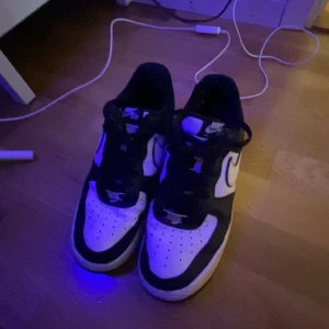 Nike Air Force 1 svartvita sneakers - Svartvita Nike Air Force 1 sneakers med klassisk design och perforerad tå. Skorna har svart ovandel med vita paneler och vit sula, samt tydlig Nike Swoosh på sidan. Skinnmaterial och snörning framtill. Perfekta för dig som gillar streetstyle.