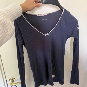 Mörkblå ribbad långärmad topp - Supersöt mörkblå ribbad långärmad topp med v-ringning och ljus spetskant samt en liten rosett framtill. Perfekt för dig som gillar en feminin och enkel stil. Mjuk och stretchig, passar till många outfits.